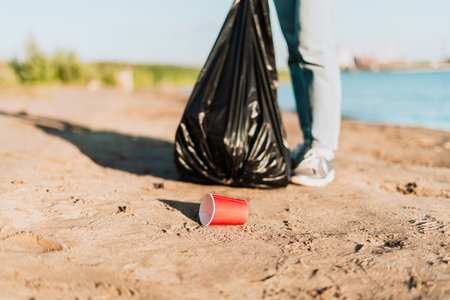 Young female volunteer picking up trash, a plastic bottles and coffee cups, clean up beach with a sea. Woman collecting garbage. Environmental ecology pollution concept. Earth Day.の写真素材