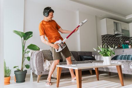 Caucasian man wearing wireless headphones plays rocks air guitar on vacuum cleaner. Male doing home...