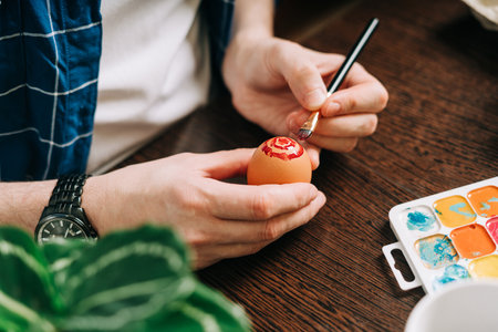 Easter day. Cropped hands of Adult man painting eggs on wooden background. Sitting in a kitchen with crayons. Preparing for Easter, creative homemade decoration.の写真素材