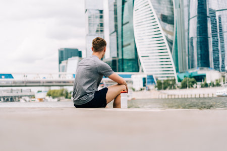 Tired Young man runner sitting on stairs and relaxing after sport training. Holding water bottle while doing workout in summer city streetの写真素材
