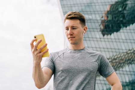 Tired Young man runner standing and using mobile phone and relaxing after sport training. Holding water bottle while doing fitness workout in city urban street, cloudy sky at summerの写真素材