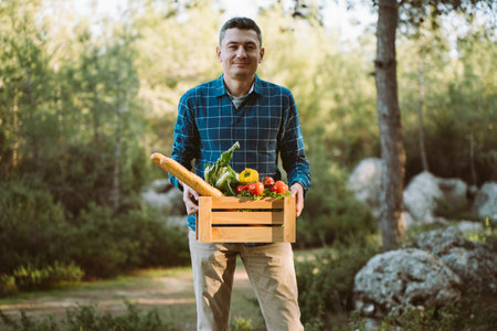 Caucasian man male holding wooden crate with vegan groceries organic vegetables and bread baguette. Farmer delivered a framed box with supplies to a farmers market camping site in the forest.の写真素材