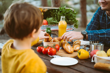 Father dad school kid boy child having a picnic in the forest camping site with vegetables, juice, coffee, and croissants. Wooden crate with fresh organic veggies surrounded with bread baguettesの写真素材