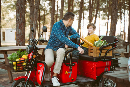 Farmer father and school boy kid delivered wooden crates with vegetables to camp site for picnic. Dad man male and son tourists delivered groceries on electric tricycle vehicle in the forrestの写真素材
