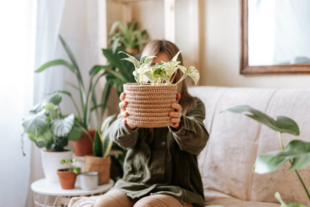 Woman holding wicker basket with plant golden pothos epipremnum, hiding face, sitting and relaxing on the couch with a lot of plants. Cozy home and urban jungle conceptの写真素材