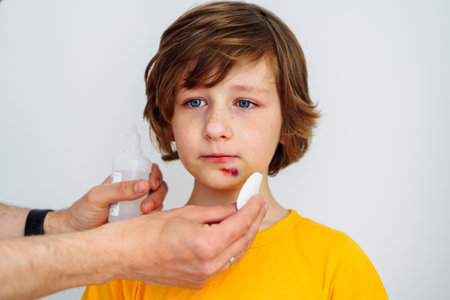 Dad doctor father treats bruised wound on his son school boy kid face. Man cleans addresses the sore wound on child face on white background with copy space for text.の写真素材