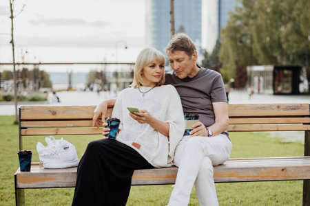 An adult mature happy couple in love sitting on bench with phone outdoors in park. A blonde caucasian man and woman spend time together and drinking coffee. Senior wife and husband walking outsideの写真素材