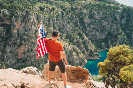 Young man standing on a rock cliff and waving the US flag while looking at sea beneath. Veteran traveller waving the American flag while standing on a mountain top. 4 fourth July Independence Dayの写真素材