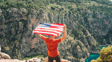 Young man standing on a rock cliff and waving the US flag while looking at sea beneath. Veteran traveller waving the American flag while standing on a mountain top. 4 fourth July Independence Dayの写真素材