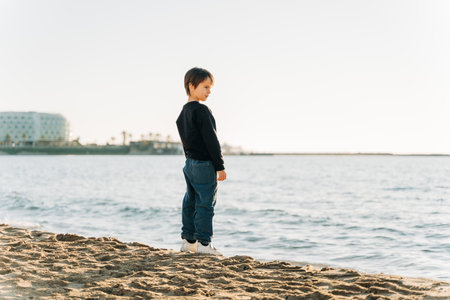 School boy child enjoying winter sea view from a sandy beach. Kid standing on shore watching autumn ocean wavesの写真素材