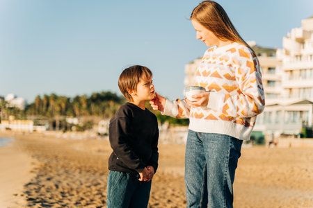 Young beautiful woman mother drinking coffee and spending free time on the winter sandy beach with her school kid son boy.の写真素材