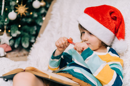 Kid boy in red Santa hat writing the letter and drawing to dear Santa at home. Child wish list. Dreams of a Christmas gifts. Merry Christmas and Happy new yearの写真素材