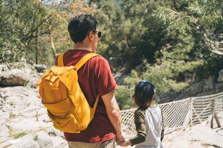 Close-up rear view of a boy and his dad walking a stone footpath in a spring forest. Child boy and father wearing casual clothes and yellow backpack while hiking in a summer greenwood forestの写真素材