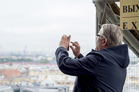 Male tourist taking pictures of European appearance on the smartphone surroundings of St. Petersburg from the colonnade of St. Isaac's Cathedral, Russia, Saint-Petersburg, September 2018.のeditorial素材