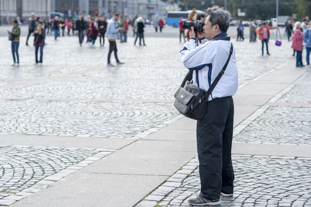 A male tourist of Asian appearance in a white jacket photographs the sights on the Palace square of St. Petersburg, Russia, September 2018. Side view.のeditorial素材