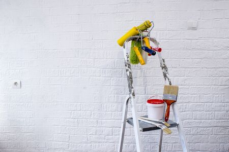 Painted metal stepladder with a bucket of paint, a set of colorful rollers and brushes on the background of a white wall decorated with bricks.の写真素材