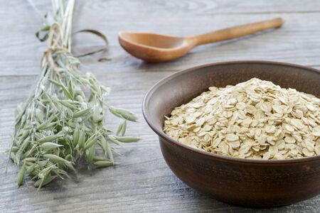 Oat flakes in vintage brown ceramic bowl with oat ears and wooden spoon on grey wooden background.の写真素材