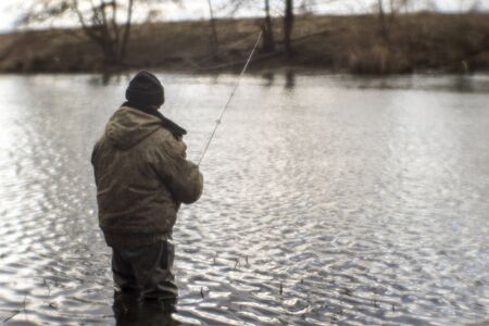 Blurred. A male angler standing in the water catches predatory fish in a cold river in the off-season. The view from the back. Background.の写真素材