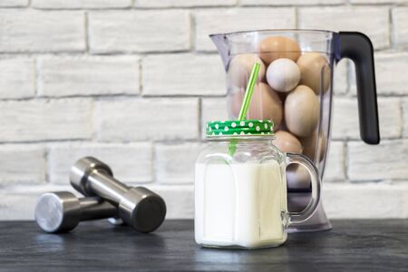 White protein cocktail in a mug on the background of a blender bowl with chicken eggs and dumbbells on a building brick wall background. Background. Concept of sports nutrition and protein diet.の写真素材