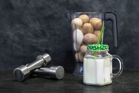 Protein shake in a mug on the background of a blender bowl with chicken eggs and dumbbells on a dark background. Background. Concept of sports nutrition and protein diet.の写真素材