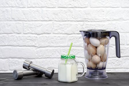 White protein cocktail in a mug on the background of a blender bowl with chicken eggs and dumbbells on a building brick wall background. Background. Concept of sports nutrition and protein diet.の写真素材