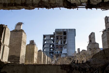 View through a gap in the concrete wall with protruding rebar on the remains of concrete Foundation piles against the background of a destroyed building in the distance and a blue sky. Background.の写真素材