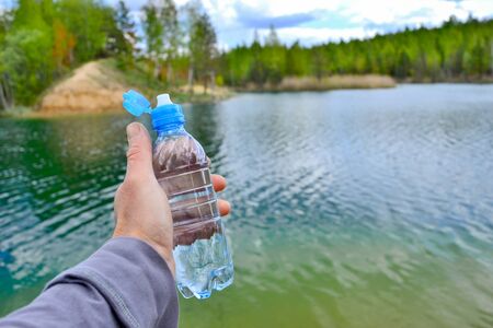 A mans hand holds a water bottle close-up against the background of clear water of a lake with a turquoise hue with the shore in the distance.の写真素材