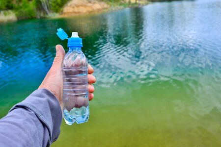A mans hand holds a water bottle close-up against the background of clear water of a lake with a turquoise hue with the shore in the distance.の写真素材