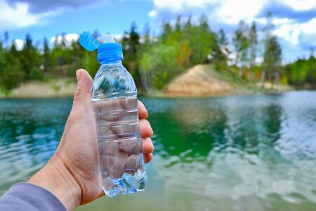 A mans hand holds a water bottle close-up against the background of clear water of the lake with a turquoise hue and a summer landscape.の写真素材