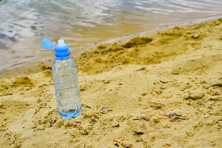 An open plastic water bottle with a close-up stands on a sandy beach against the background of clear lake water.の写真素材