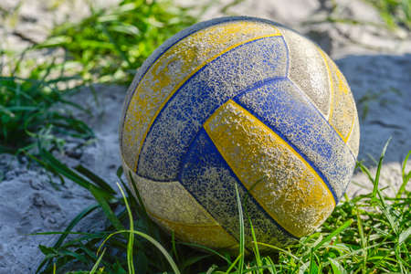 The yellow-blue volleyball ball is old, scratched lying on the sand at the edge of the grass, illuminated by the evening sunの写真素材