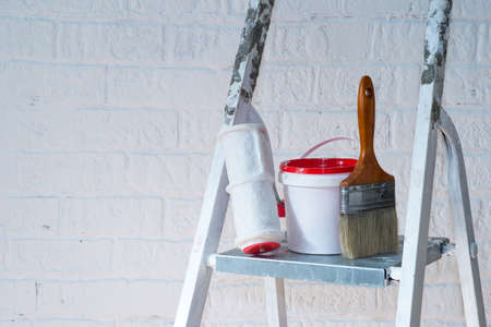 A brush, bucket and textured roller under a brick stand on a metal stepladder near a white wall decorated with bricks.の写真素材