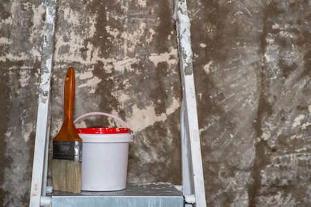 A paintbrush and bucket with a red lid stand on a metal stepladder on the background of an concrete...