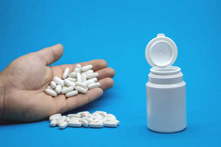 A man's hand with a bunch of pills and an open white bottle on a blue background. background.の写真素材
