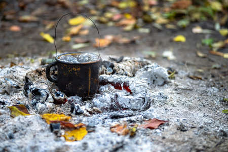 Metal black smoked mug with boiling water on the coals of a fire on the background of autumn leaves. Background.の写真素材