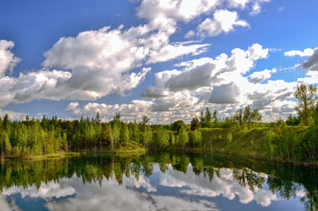 Lake with banks in the bright spring green of the forest with a reflection of the blue sky with clouds in the water surface.の写真素材