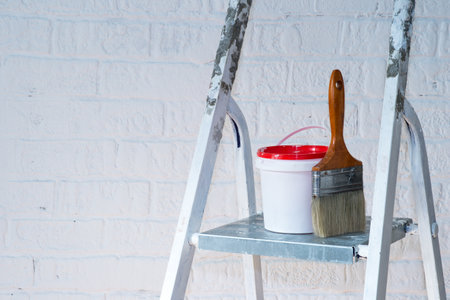 A paintbrush and bucket with a red lid stand on a metal stepladder against a white wall decorated with bricks.の写真素材