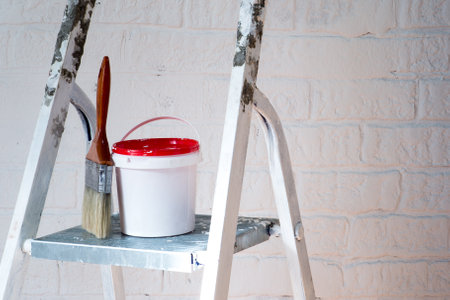 A paintbrush and bucket with a red lid stand on a metal stepladder against a white wall decorated with bricks.の写真素材