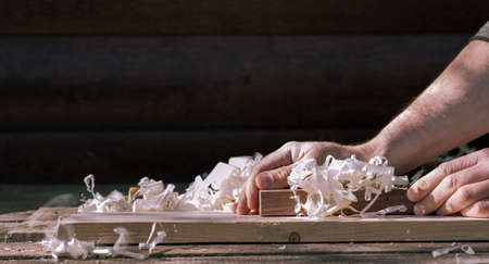 Men's hands close-up planning a board on a workbench with a pile of shavings on the background of a dark wall of the house with a Japanese wooden plane. The concept of finishing wood processing.の写真素材
