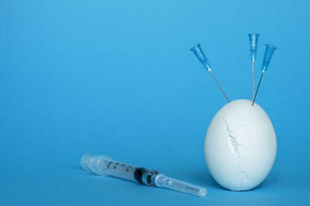 A syringe and a white egg in cracks with needles on a blue background. The concept of immunity risk in vaccinated youth.の写真素材
