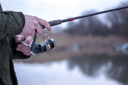 The frozen hands of a male angler close-up to hold the spinning in a blurry motion wind the fishing line on a reel against the background of a winter river. Selective focus on the hand. background.の写真素材