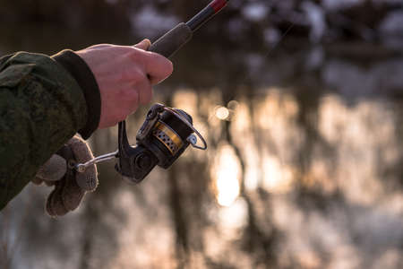 The frozen hands of a male angler holding a spinning reel wind a fishing line on a reel against the background of the river. Selective focus on rod coil. background.の写真素材
