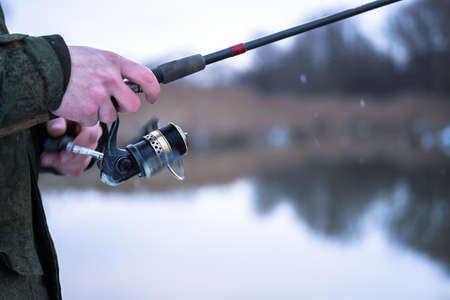 blurred. The frozen hands of a male angler close-up to hold the spinning in a blurry motion wind the fishing line on a reel against the background of a winter river and falling snowflakes. background.の写真素材