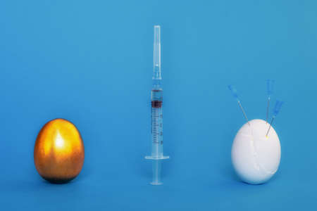 A metallic golden egg and a white chicken egg in cracks and needles from syringes and a syringe on a blue background. The concept of probable weakening of immunity in young people after vaccinations.の写真素材
