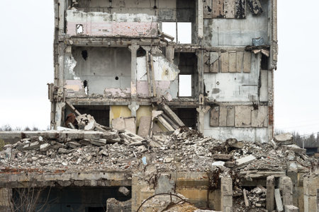 A ruined building against a gray sky with piles of concrete rubble and protruding piles in the...