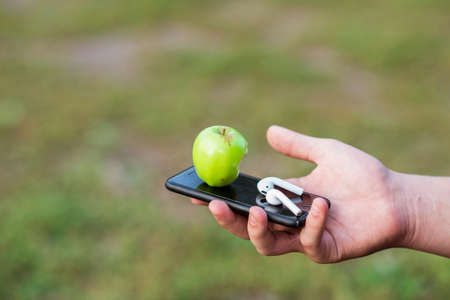 The teenagers hand holds a mobile phone with a bitten green apple on the background of a blurred meadow. Background. The concept of Internet addiction of modern lifestyleの写真素材