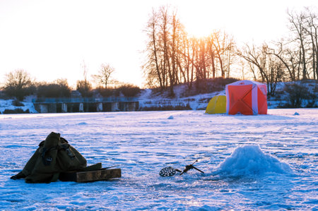 Winter fishing rod with a bent nod over a drilled hole, retro backpack on the background of a dam...