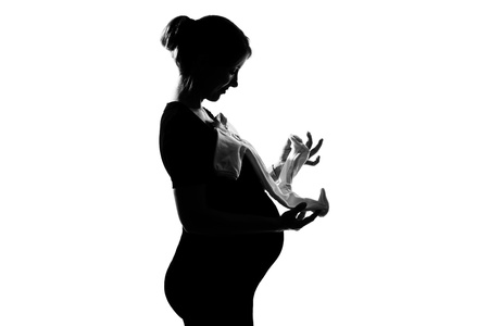 one beautiful caucasian pregnant woman holding baby clothes in silhouette on studio isolated white backgroundの写真素材