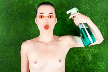 Portrait of a topless woman wearing tanning bed glasses with strawberry in mouth and spray bottle in hand against greenの写真素材