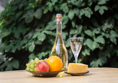 Composition of cheese, grapes, peaches, white grapes, bottles and glasses of wine on a wooden round table. in the courtyard. against the background of the vineの写真素材